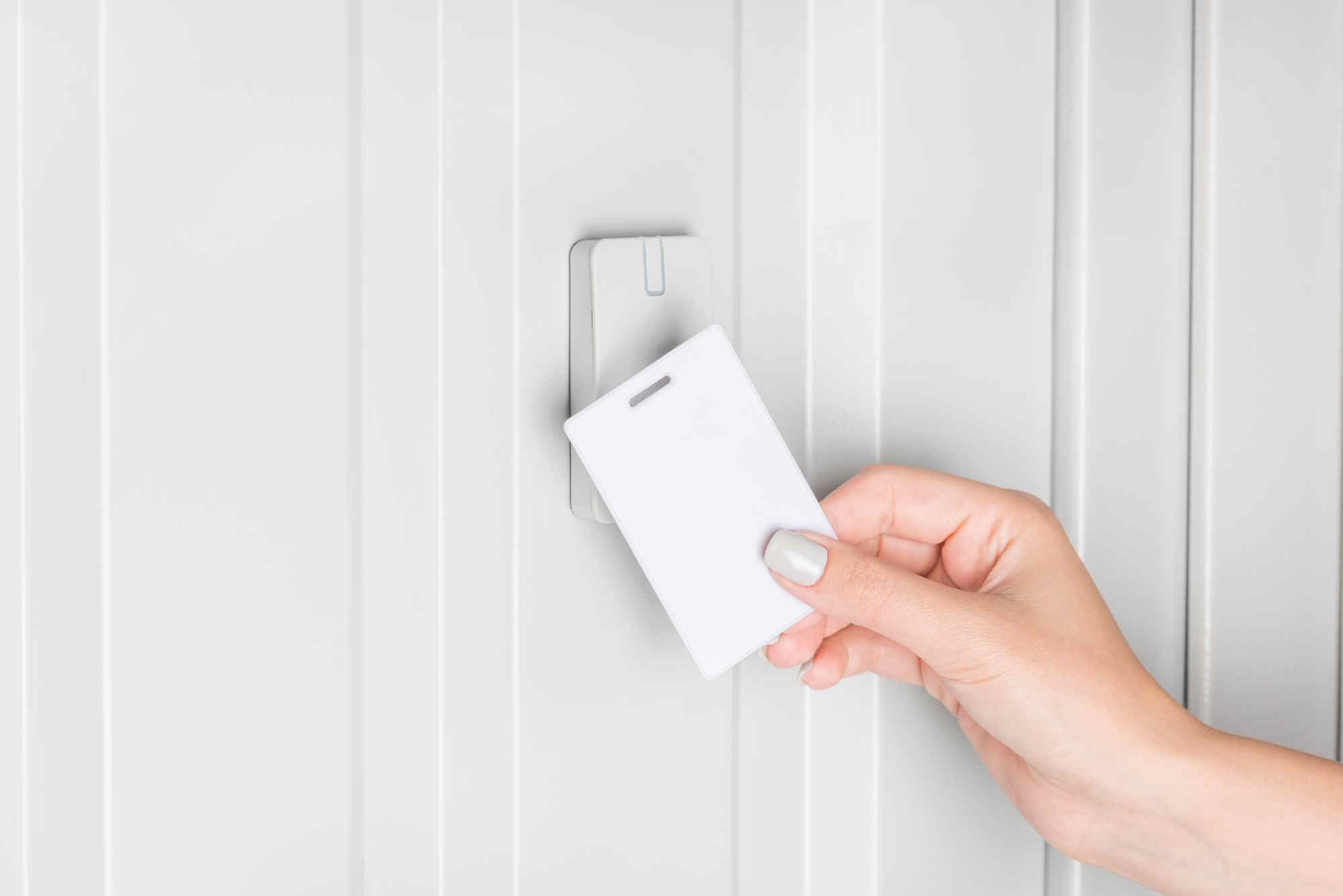 A hand holds a white key card up to an electronic card reader mounted on a white vertical-paneled wall.
