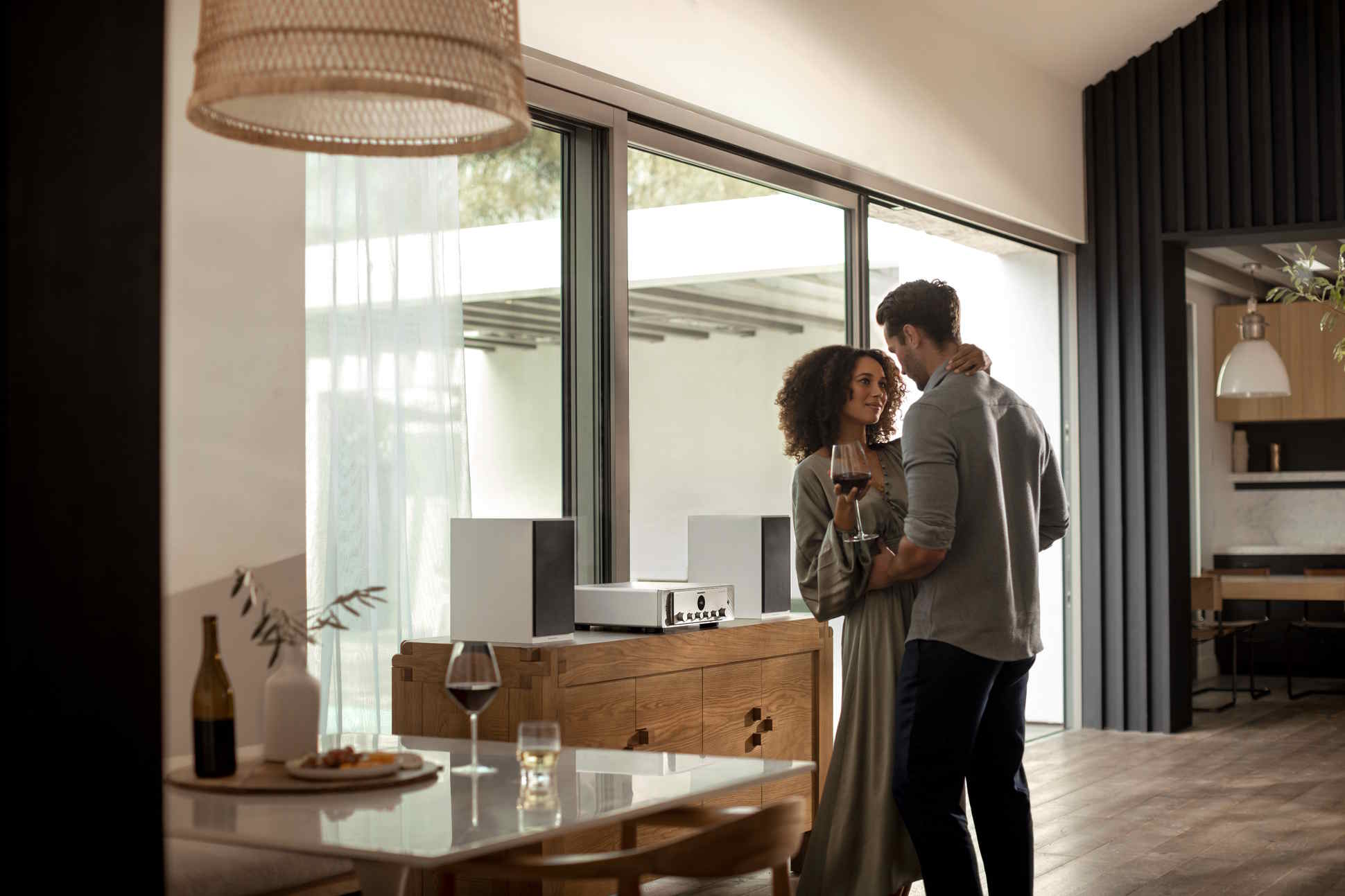 A romantic couple holds wine glasses and embraces in a luxury home next to an elegant audio system and large glass doors.