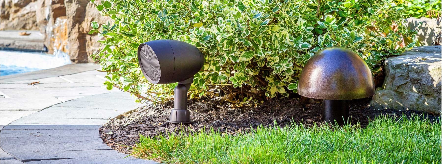 An outdoor directional speaker and a dome-shaped landscape speaker installed in a garden bed next to a stone pathway and pool.
