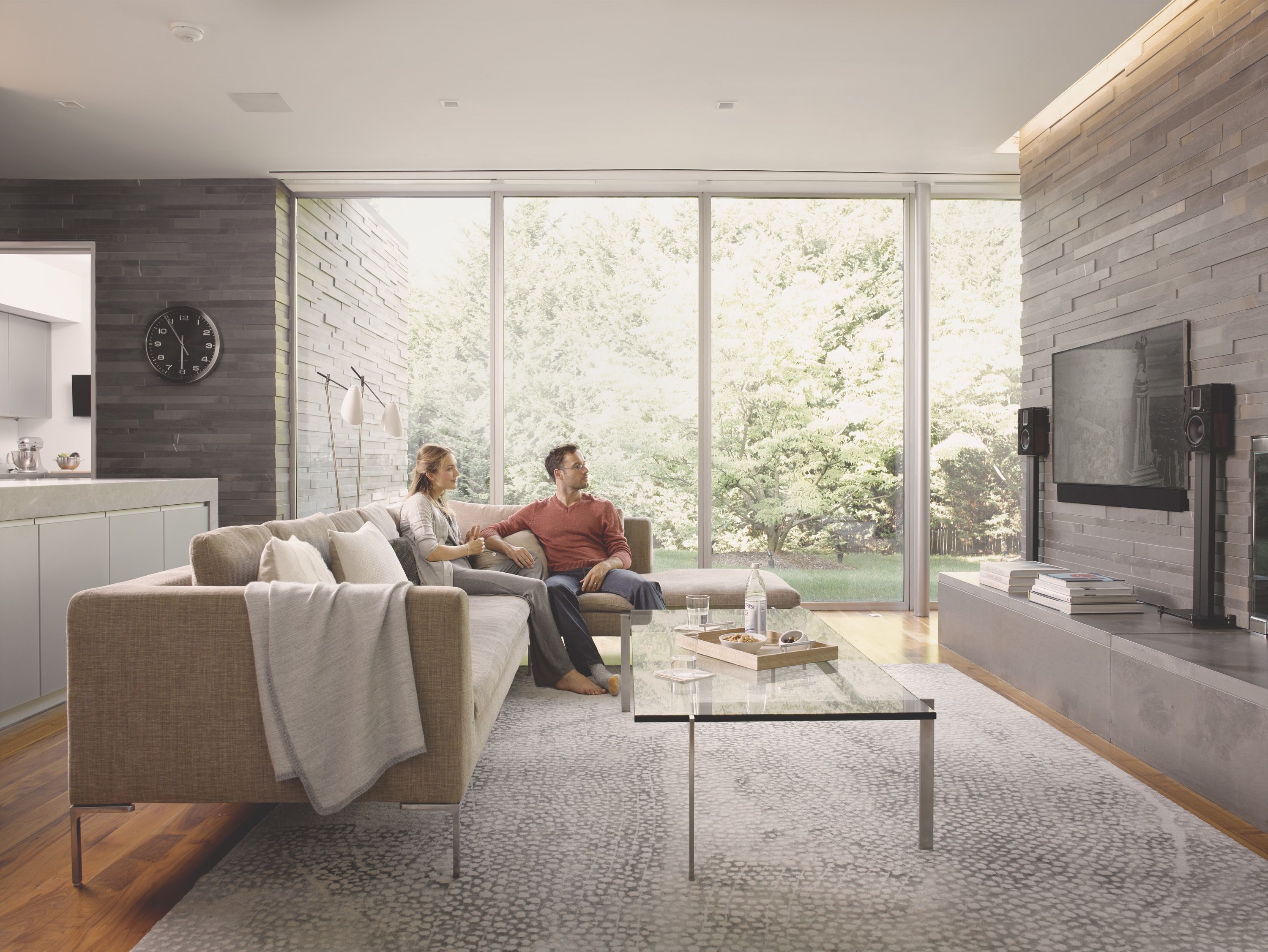 A couple sits on a modern sectional sofa in a luxury living room with stone accent walls and floor-to-ceiling windows looking out at trees.