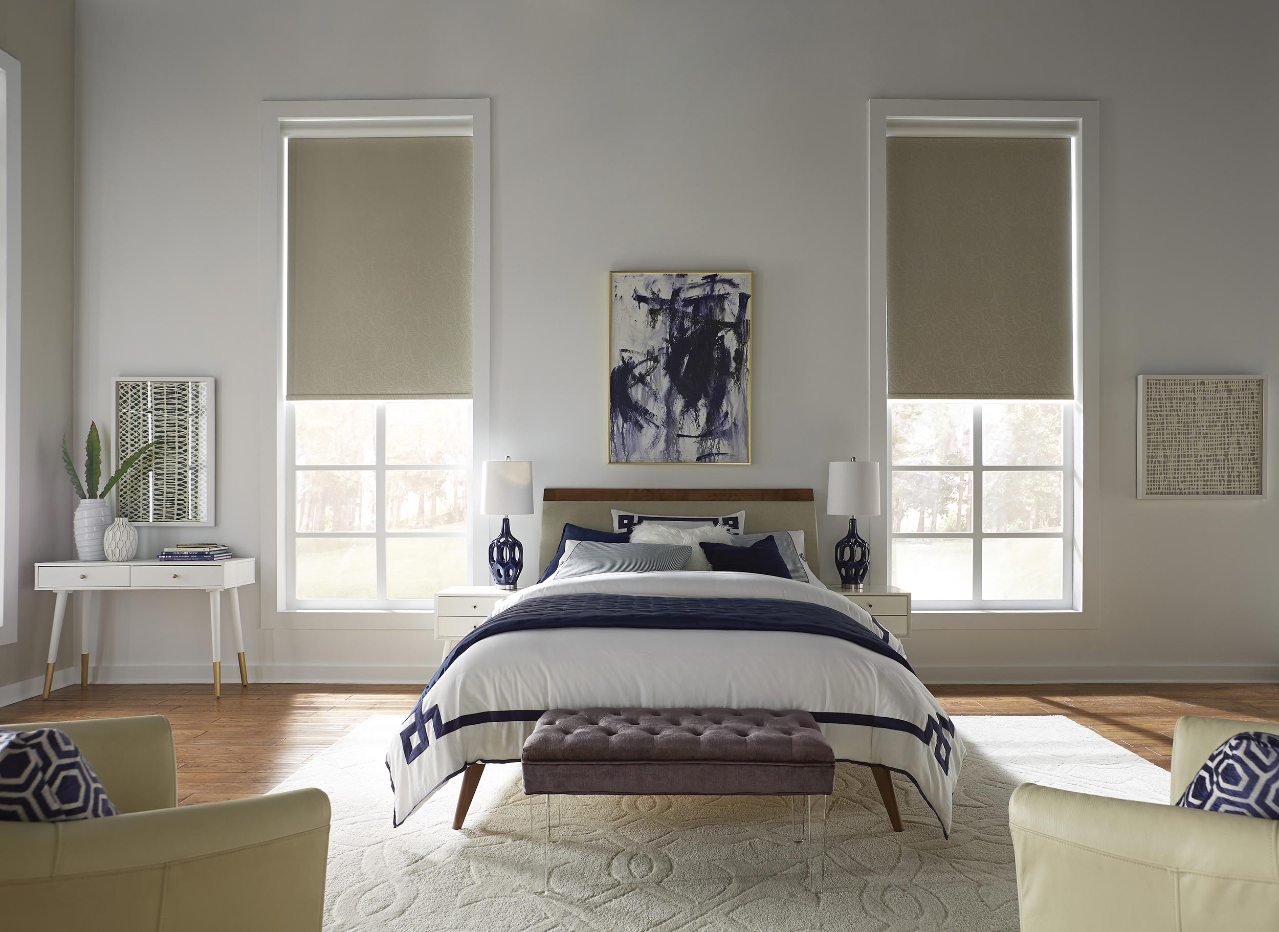 A elegant, modern bedroom featuring a bed with navy accents, abstract wall art, and two large windows with beige roller shades.