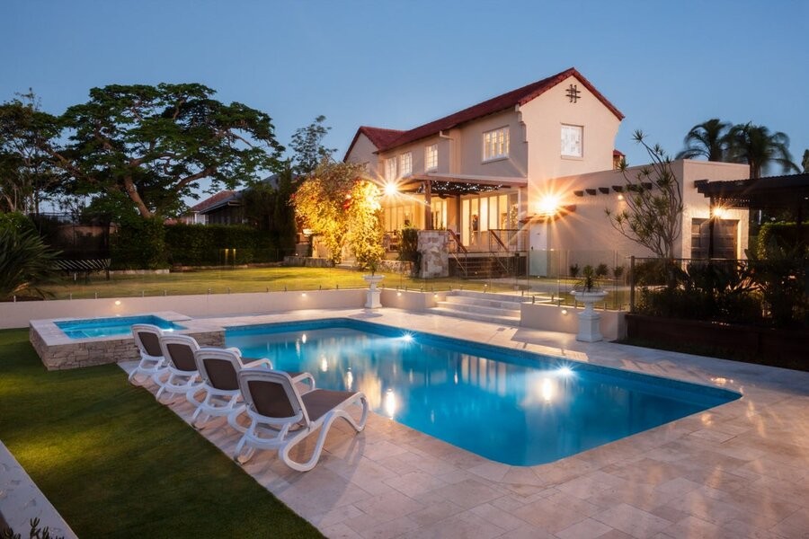 A backyard featuring a pool and exterior lighting with a house in the background.