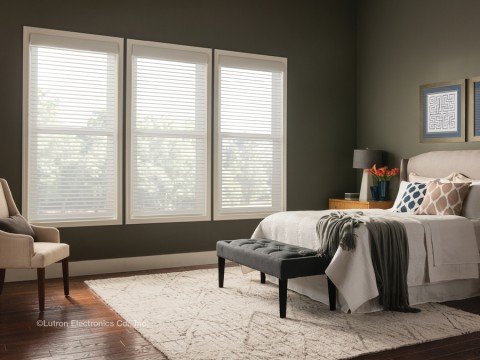 A bedroom with a bed with blankets draped over it and a chair facing it with light filtering in through sheer shades. 