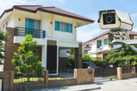 A security camera is prominently mounted in the foreground, monitoring a modern suburban home with a fenced yard, trees, and a driveway.